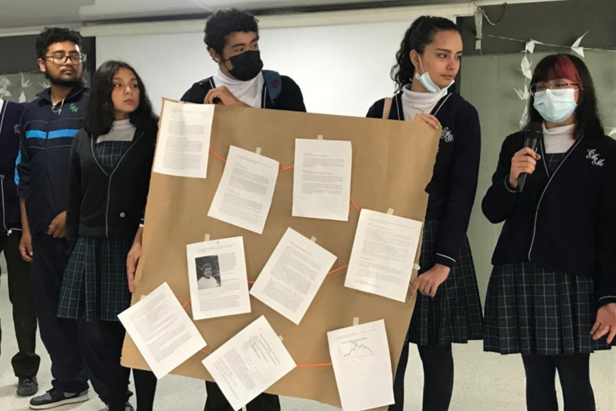 Foto de un grupo de 5 estudiantes exponiendo una cartelera con varias hojas pegadas allí 