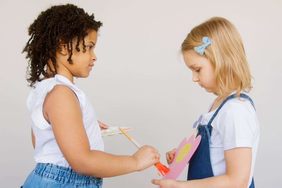 Dos niñas de frente una está pintando una hoja que tiene la otra niña en las manos