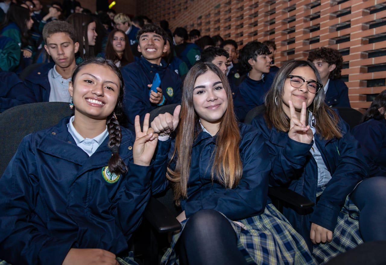 Foto de 3 estudiantes alegres en un auditorio con más compañeros atrás 